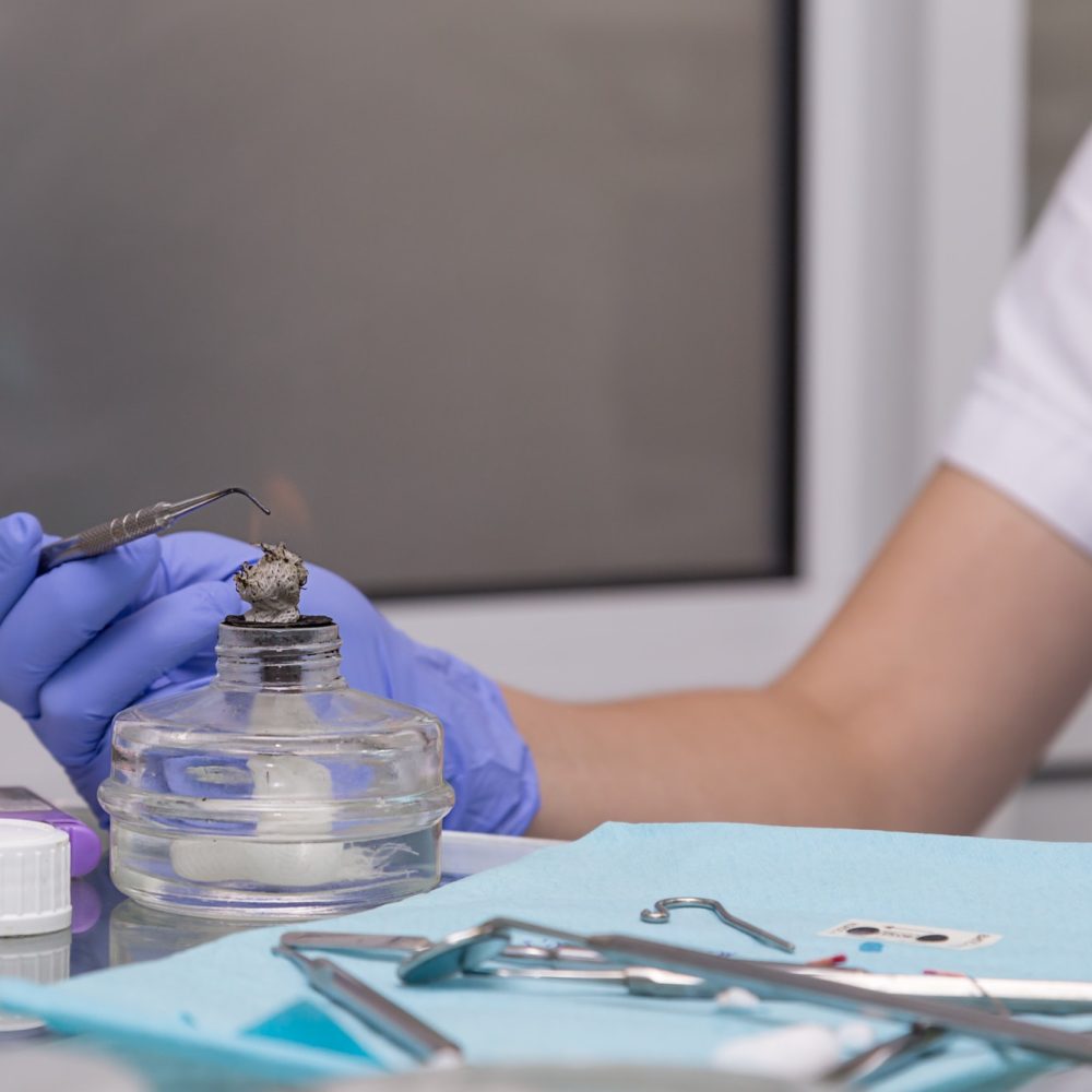 Mano con guante azul sosteniendo un instrumento dental sobre un frasco con material, en un entorno de laboratorio dental. Se observan herramientas y suministros médicos en la mesa.